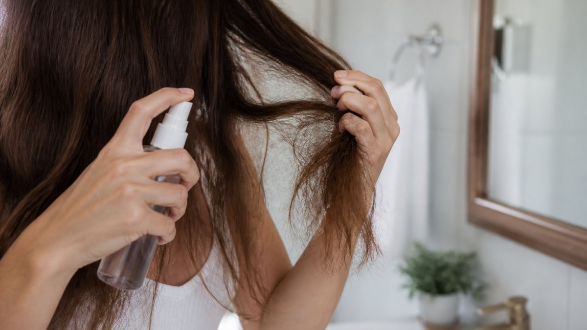 A close-up of a woman's hands applying a leave-in conditioner spray to her dry, brittle hair ends in a bright bathroom setting