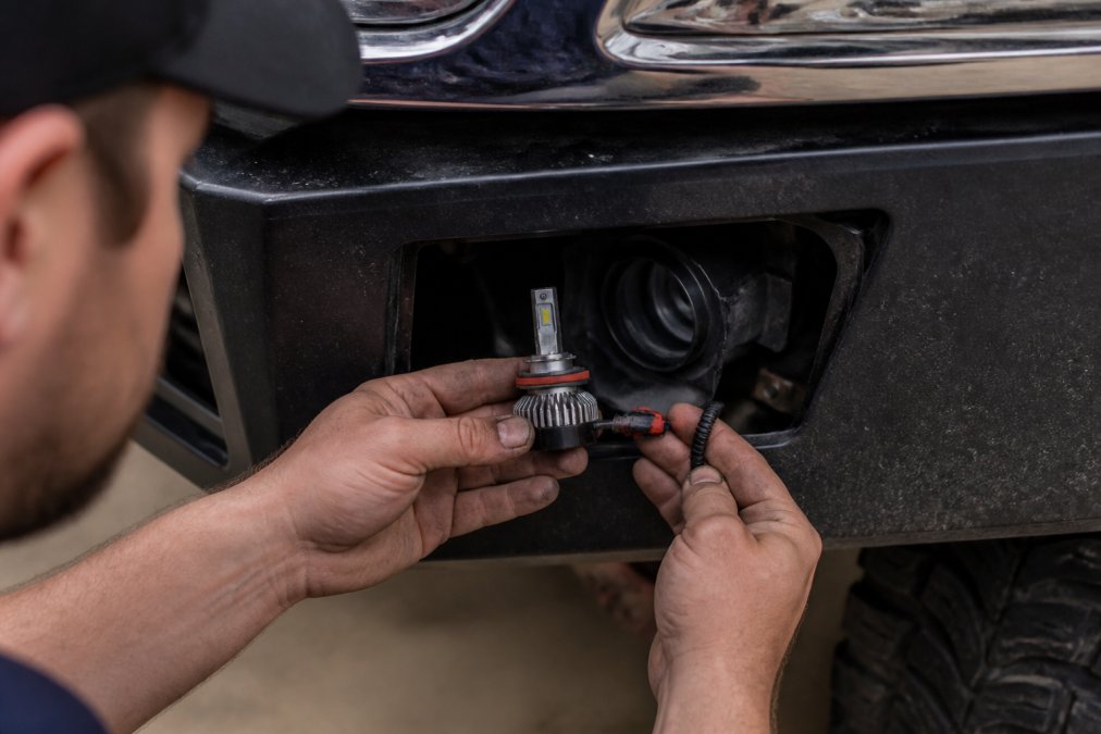 Mechanic's hands carefully installing an LED fog light bulb into a truck's fog light housing, showing the wiring harness connector and mounting bracket in sharp focus