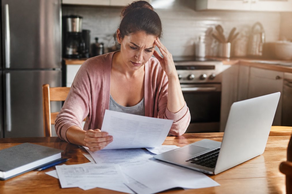 A person sitting at a kitchen table reviewing legal documents and court papers with a concerned expression, a laptop and pen nearby in warm afternoon light