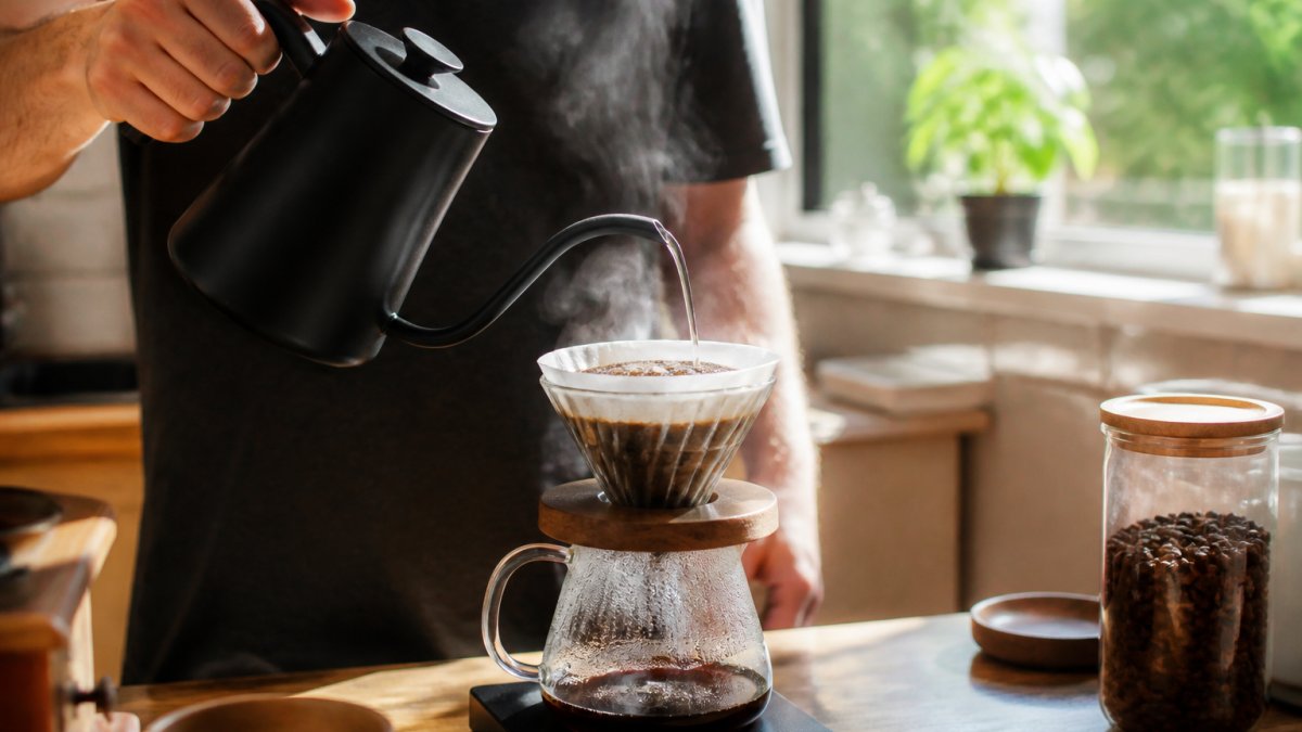 Hands pouring hot water in a steady spiral over light roast coffee grounds in a V60 dripper, with steam rising during the bloom phase, natural morning light streaming through the kitchen window