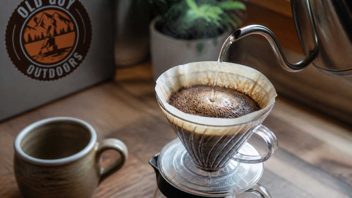 A close-up of a pour-over cone with a gooseneck kettle pouring hot water over light roast coffee grounds, steam rising, on a wooden kitchen counter with a mug nearby