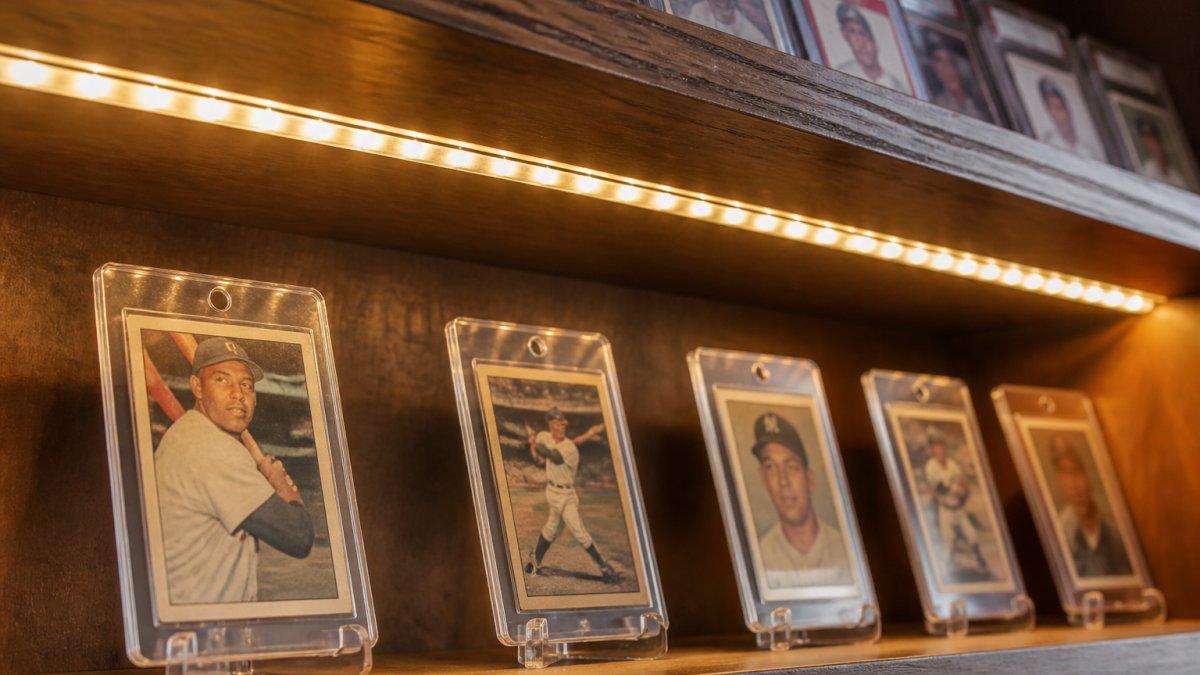 Close-up of a warm white LED strip glowing along the underside of a wooden shelf holding baseball cards in protective cases, soft light pooling on the cards below