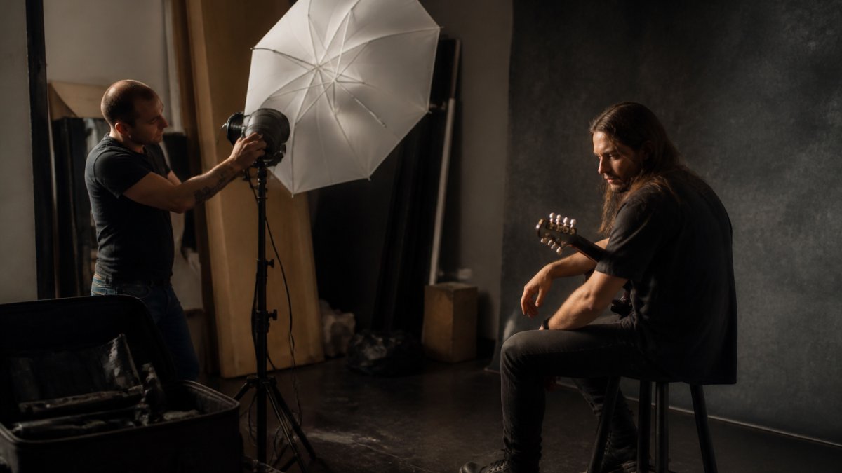 A photographer adjusting a single strobe with a large umbrella modifier on a light stand in a dark studio, with a musician seated on a stool facing slightly away from the light source, creating deep shadows across one side of their face.
