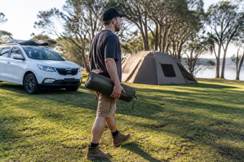 A person easily carrying a compact folded camping chair under one arm while walking across a grassy Australian campsite with a car and tent in the background
