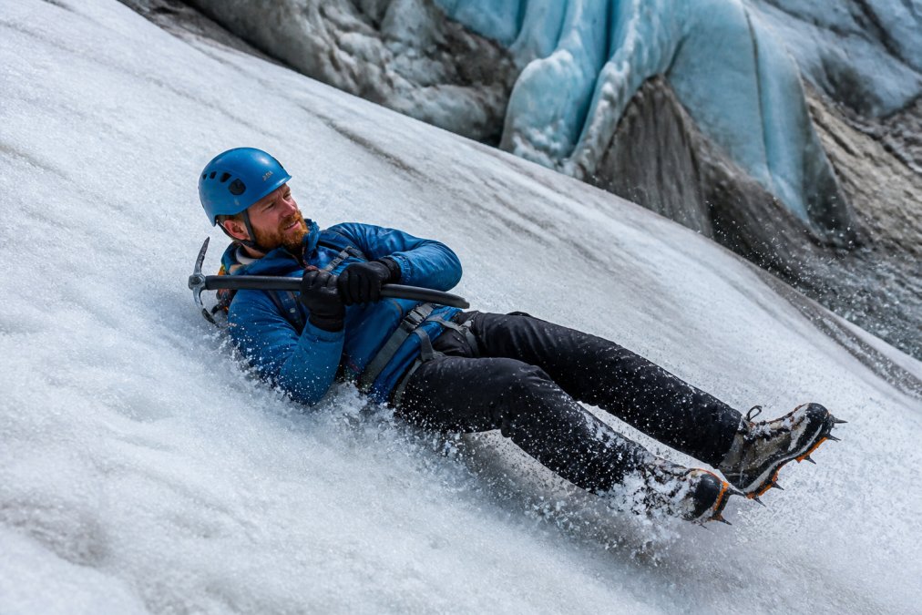 Climber demonstrating self-arrest technique on snowy glacier slope with proper body position, axe placement across chest, crampons dug in, and ice background showing dynamic stopping motion