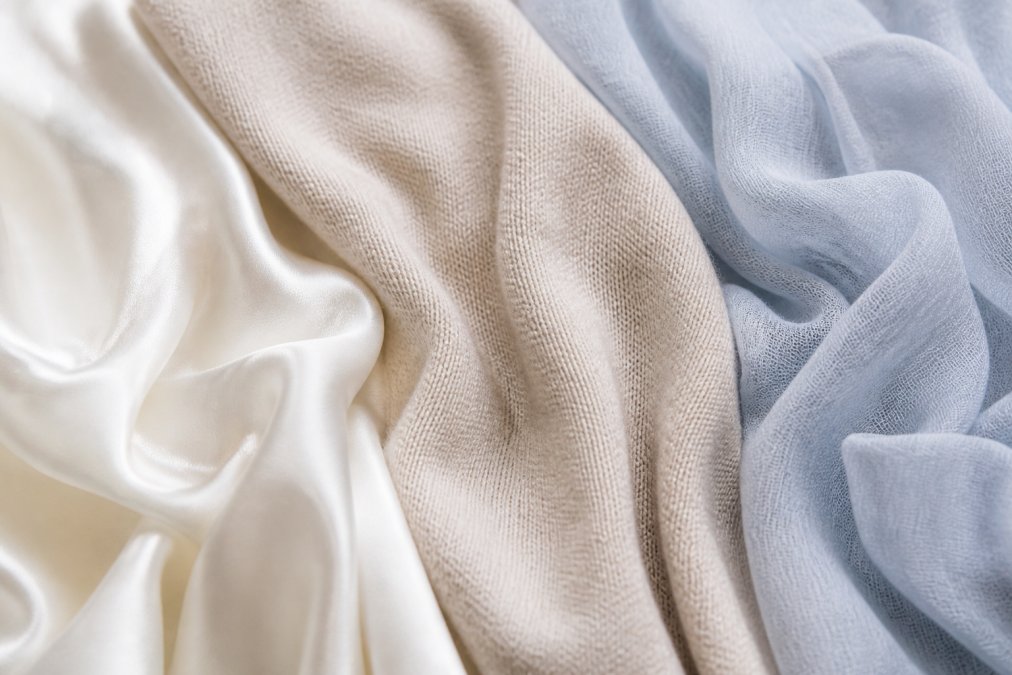 Close-up of lightweight fabric textures showing silk, cashmere, and modal blends laid flat, displaying the delicate weave and breathable quality of each material in natural daylight