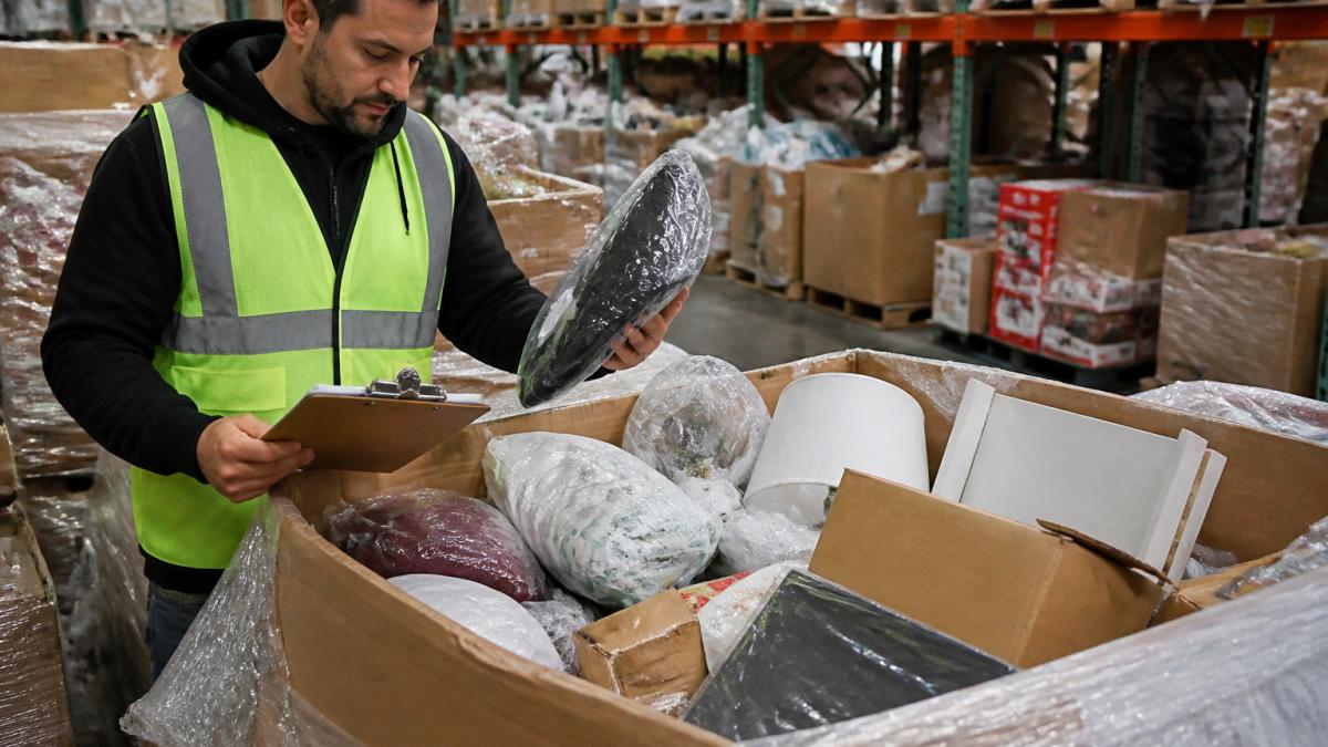 A reseller in a warehouse opening a liquidation pallet, sorting through mixed customer returns and overstock inventory with a clipboard and manifest in hand