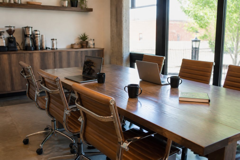A conference table in a bright, modern meeting room with comfortable seating, laptops open, and a coffee station visible in the background, natural light from large windows