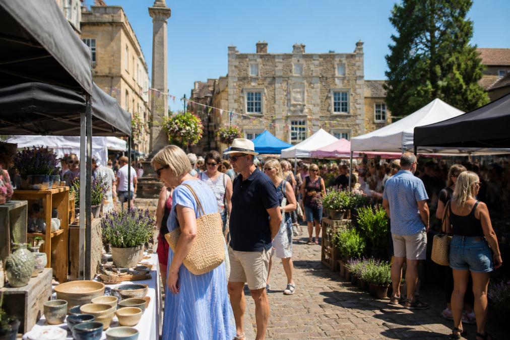 Visitors browsing handmade craft and food stalls at an outdoor artisan market in a sunlit historic town square, with a traditional stone building and bunting visible in the background