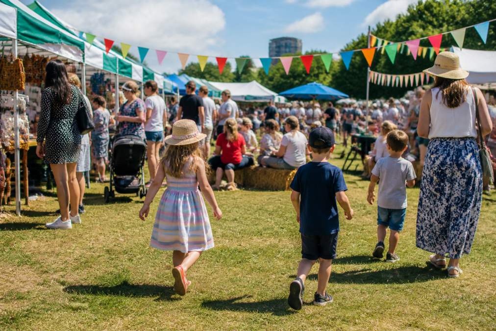Families with young children enjoying an outdoor community event on a sunny afternoon, with colourful market stalls, bunting, and a grassy open space in the background