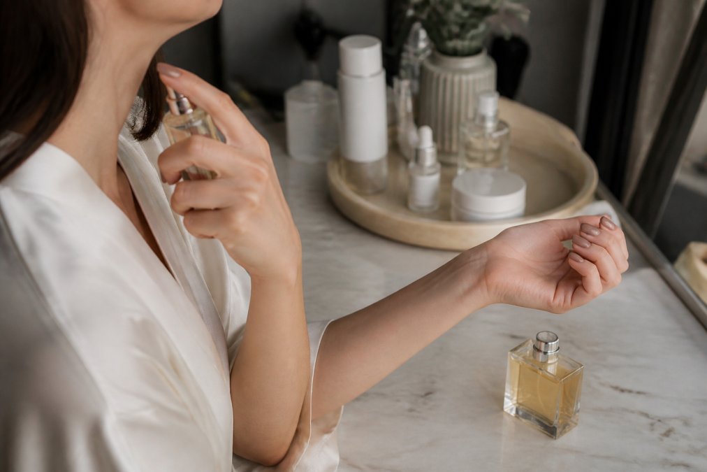Hands applying perfume to pulse points on wrist and neck, with fragrance bottle nearby on vanity with skincare products