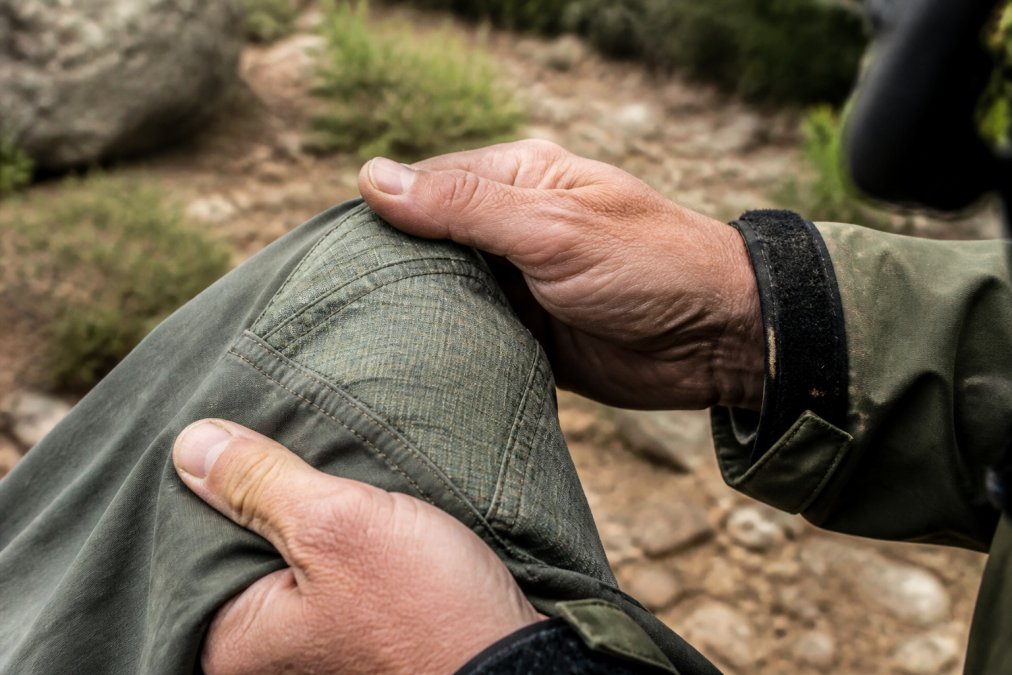 Weathered hands inspect reinforced stitching on durable outdoor adventure clothing during a mountain hike.