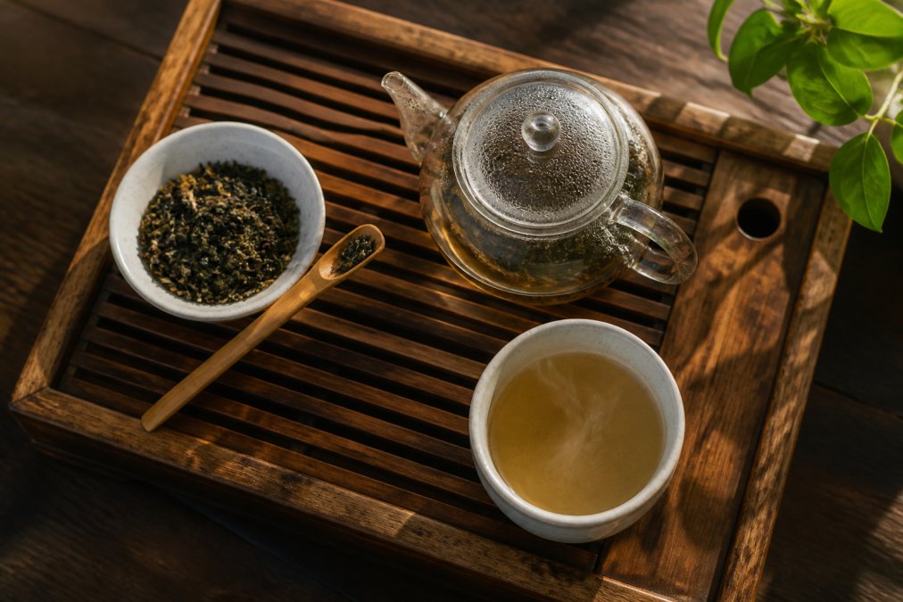 Glass teapot and bowl of loose leaf tea on a wooden tray with a bamboo scoop and steaming ceramic cup