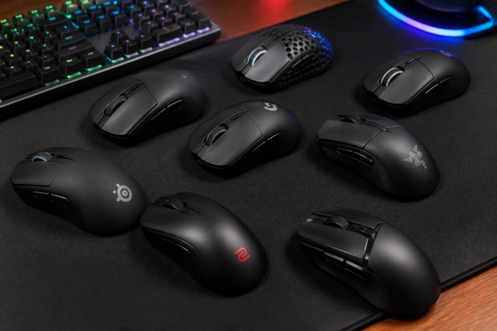 Close-up flat-lay of multiple gaming mice arranged on a dark gaming mousepad, with a mechanical keyboard and RGB lighting casting colored light across the desk surface