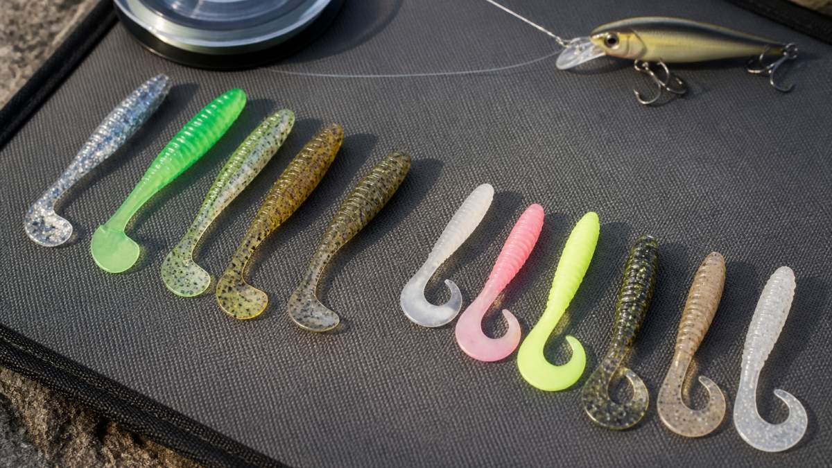 Close-up of soft plastic swimbaits and grubs arranged on a tackle mat showing multiple colors and tail designs, with fishing line and a walleye lure in natural daylight