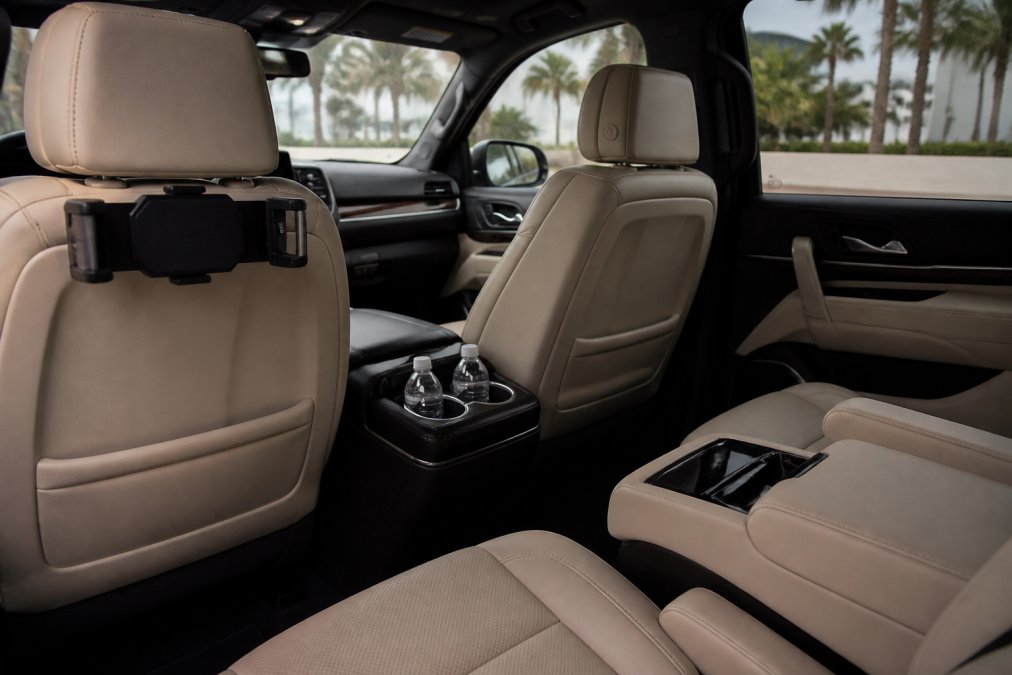 Interior view of a luxury black SUV with cream leather seats, soft ambient lighting, chilled bottled water in the center console, and a tablet mount on the back of the front seat, photographed from the rear passenger perspective