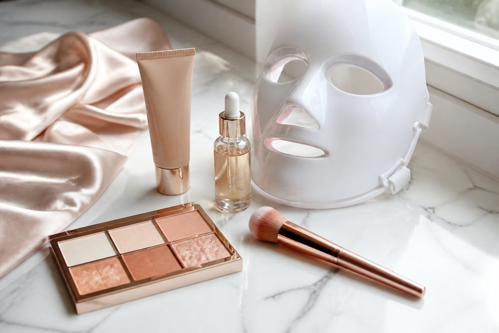 A flat-lay photo of five luxury beauty products arranged on a white marble vanity counter, including a tinted moisturizer tube, a glass serum dropper bottle, a large face palette, a rose gold brush, and a white LED mask, styled with a silk scarf and soft morning window light