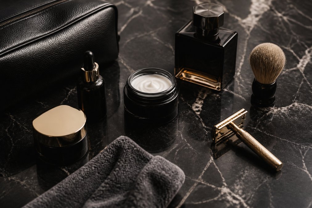 An elegant flat lay of premium men's grooming products including a skincare set, a cologne bottle, and a high-end grooming tool on a dark marble surface with soft, dramatic lighting