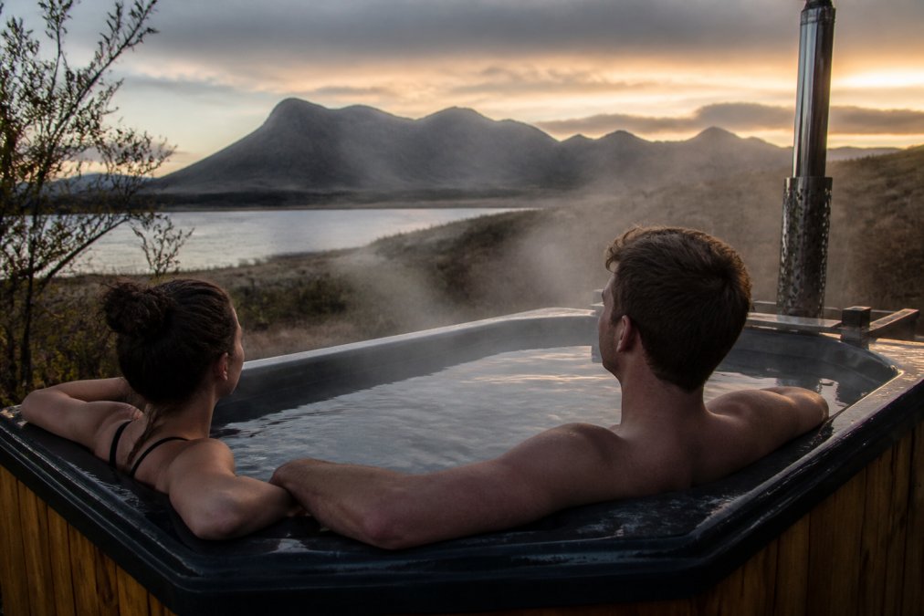 Couple relaxing in a private outdoor hot tub at dusk, with Scottish Highland mountains visible across misty terrain and warm steam rising into cool evening air