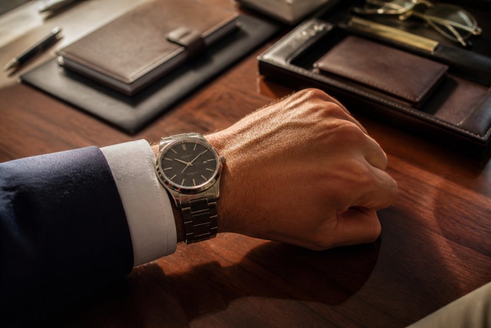 Close-up of a man's wrist wearing an engraved luxury watch, refined office desk with leather accessories and warm afternoon light streaming across polished wood
