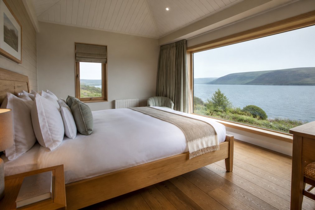 A luxurious lodge bedroom with a handmade bed, crisp linens, and large windows framing a tranquil loch view on the Isle of Skye