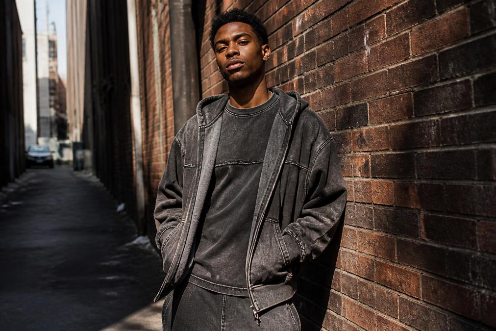 A confident young man wearing an oversized snow-washed zip hoodie and raw-seam boxy tee, leaning against a weathered brick wall in a narrow urban alley, moody late-afternoon city light casting strong shadows across the textured fabric