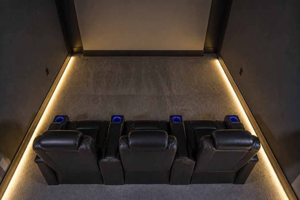 Overhead view of a compact rectangular room with three dark leather theater recliners arranged in a single row close to the back wall, a clear walkway of roughly three feet visible between the front of the seats and the screen wall, ambient lighting strips along the baseboards