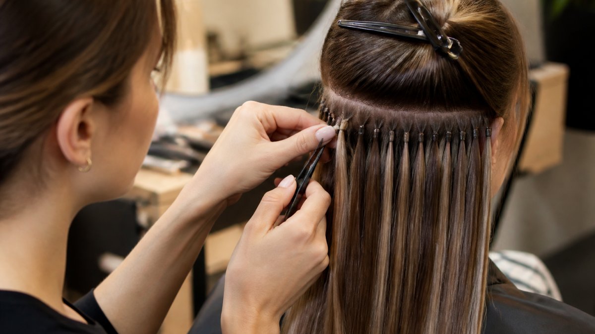 Professional stylist carefully applying nano-bonded hair extensions to client's roots, showing seamless blend between natural and extension hair with precision placement, warm salon lighting highlighting the cuticle alignment