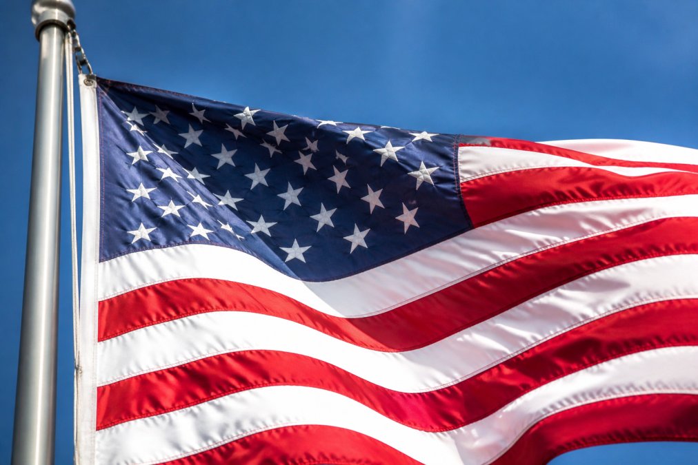 Close-up of a high-quality American flag flying on a steel flagpole outdoors, showing clearly embroidered stars and individually sewn stripes in vivid red, white, and blue against a bright blue sky with sunlight catching the fabric