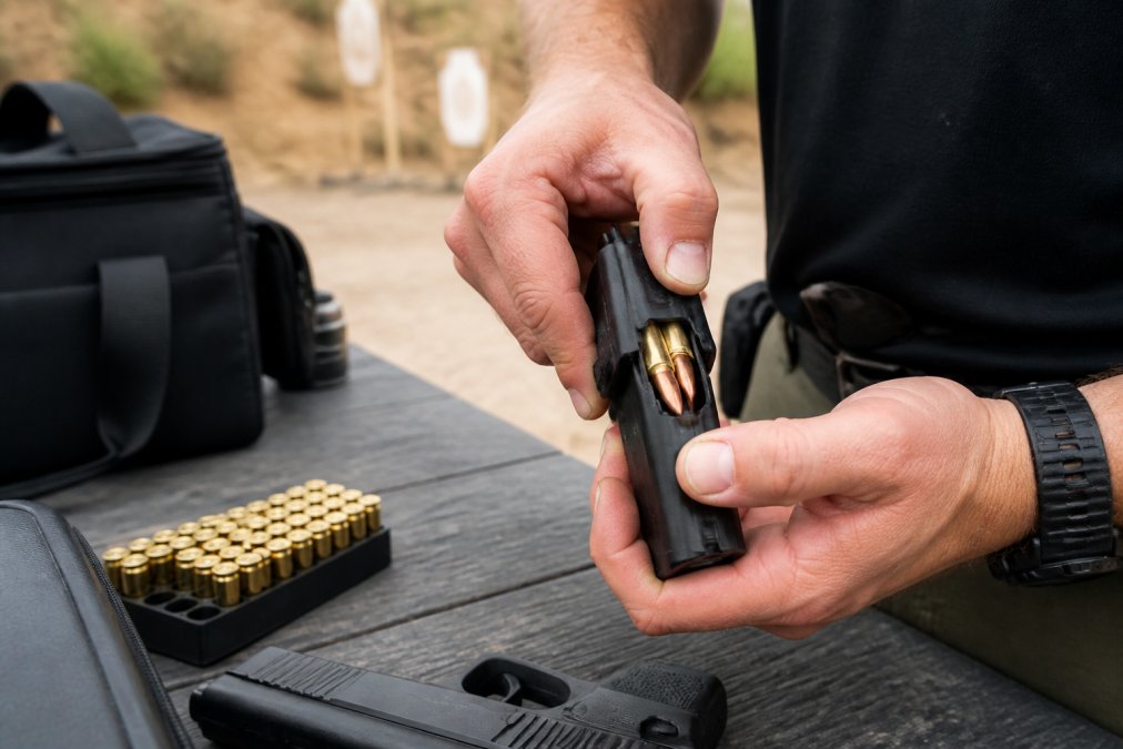 Close-up of hands demonstrating proper grip and positioning while inserting a 9mm magazine into a speed loader at a shooting range, natural outdoor lighting with ammunition and range bag visible