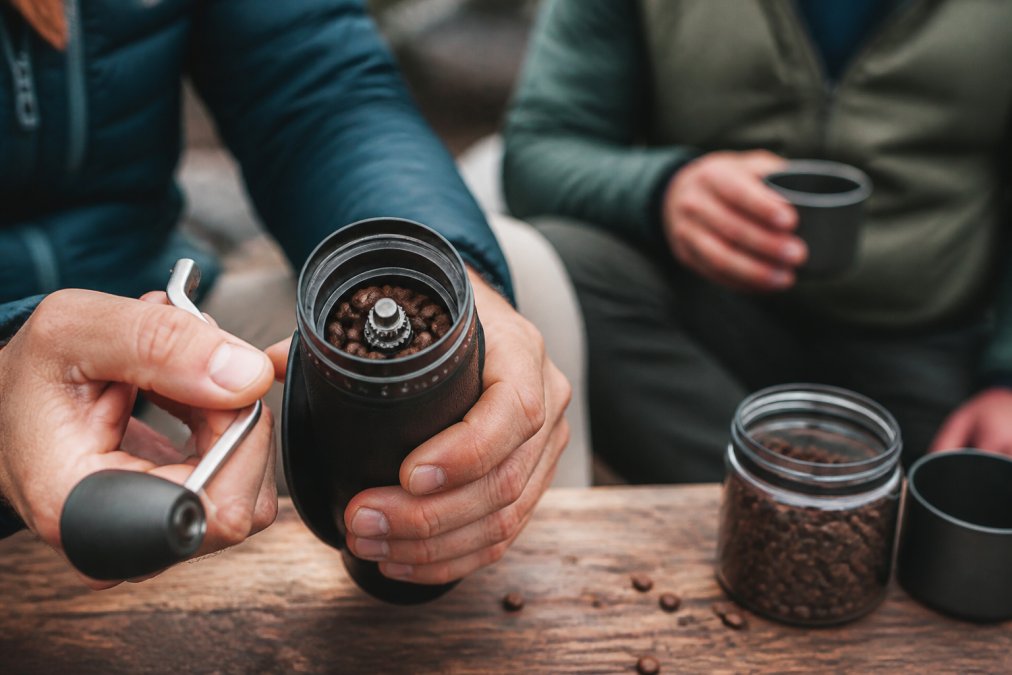 Close-up of hands holding a manual coffee grinder with visible burr mechanism and adjustment ring, beans visible in the hopper, natural lighting