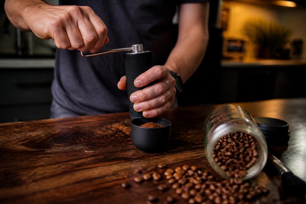 Hands holding a manual coffee grinder over fresh grounds on a dark wooden kitchen countertop