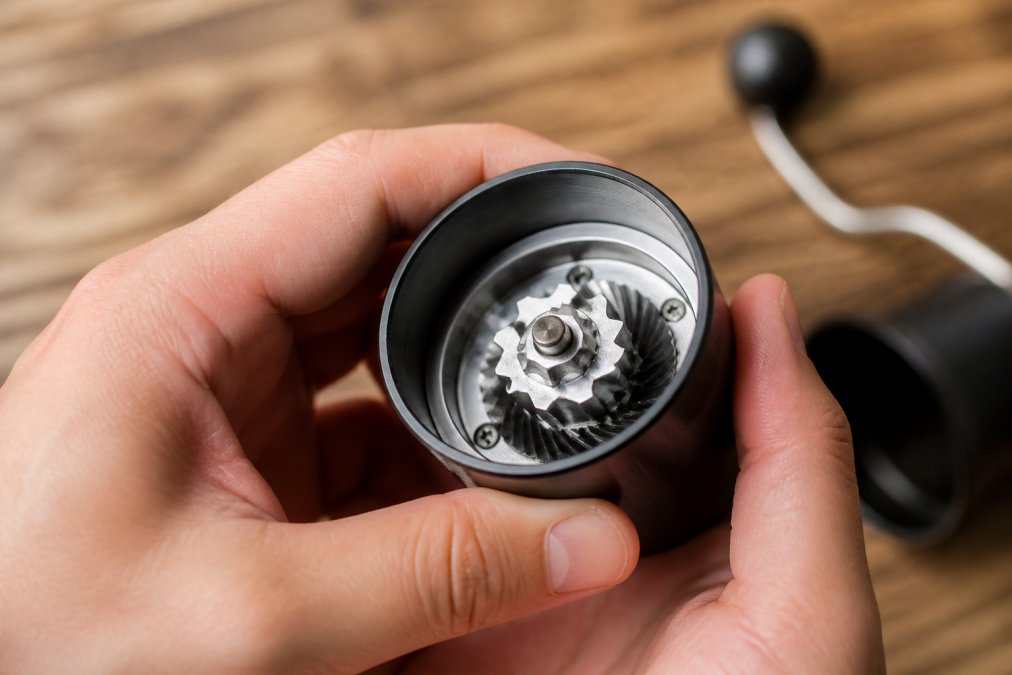 Close-up of hands holding a manual coffee grinder with visible burr geometry inside, showing the internal grinding mechanism with stainless steel burrs catching light