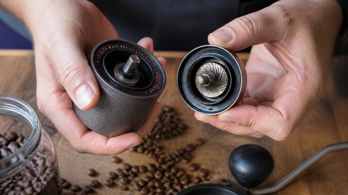 Close-up of hands holding a manual coffee grinder with whole beans scattered nearby, showing the adjustment dial and conical burr mechanism clearly visible