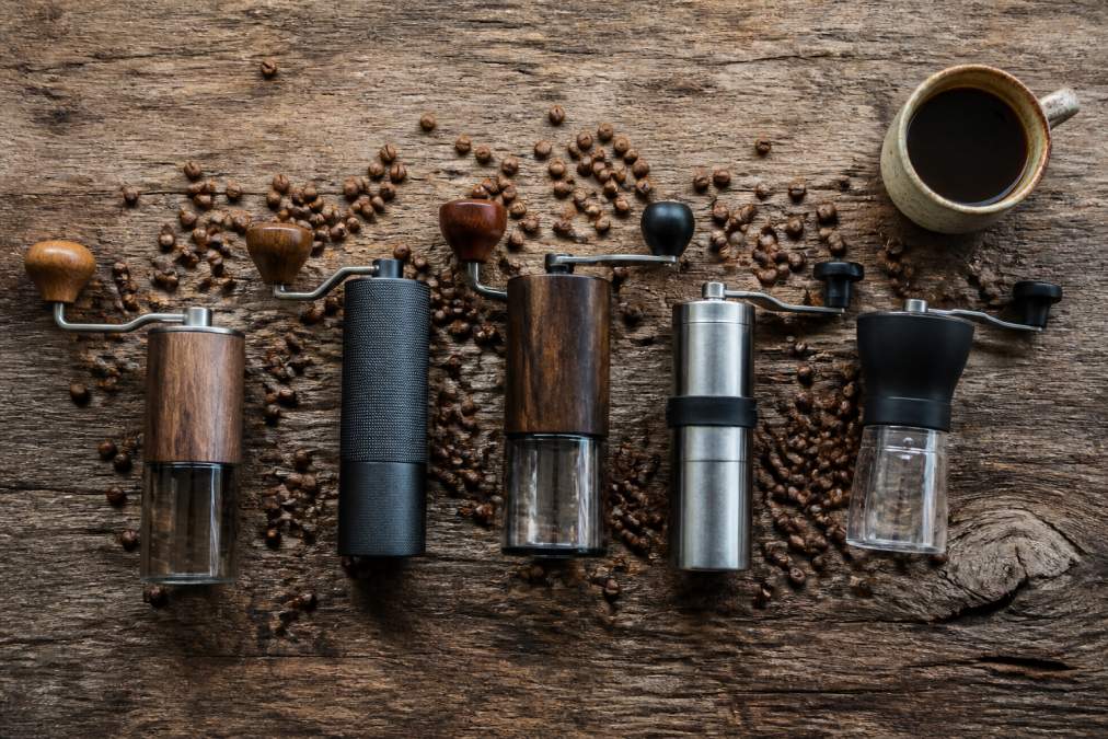 Flat lay arrangement of five different manual coffee grinders, Comandante, 1Zpresso, Timemore, Porlex, and Hario, arranged on a weathered wooden surface with freshly roasted coffee beans scattered around them and a small ceramic cup in the corner