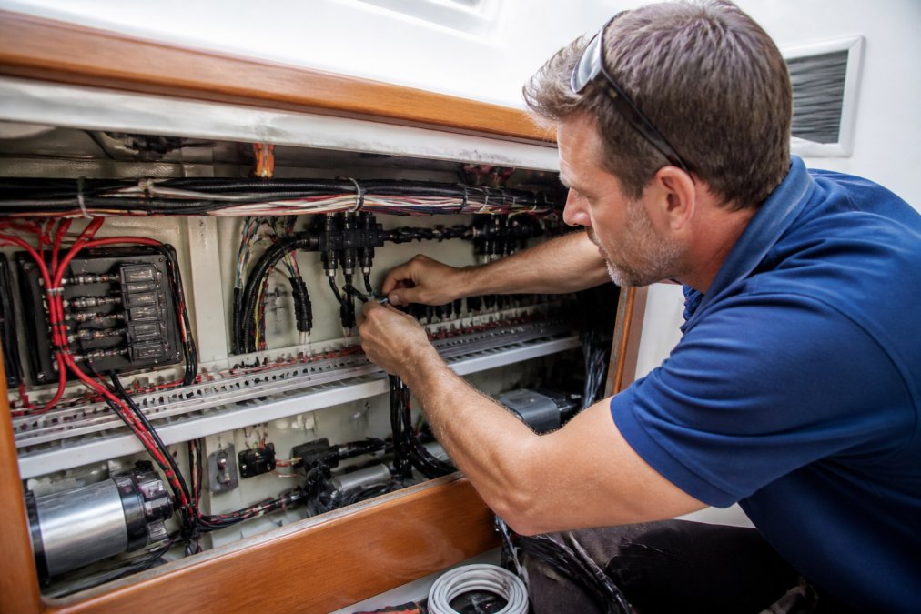 A marine technician in a blue work shirt routing NMEA 2000 cables through an open electrical panel below deck on a sailboat, with wiring harnesses, T-connectors, and a cable tray visible in a clean, well-lit installation
