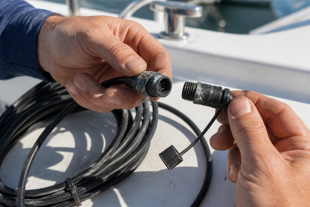 A boat owner inspecting a marine network cable for corrosion damage near the connection point, with visible salt spray residue and weathered cable jacket in bright sunlight