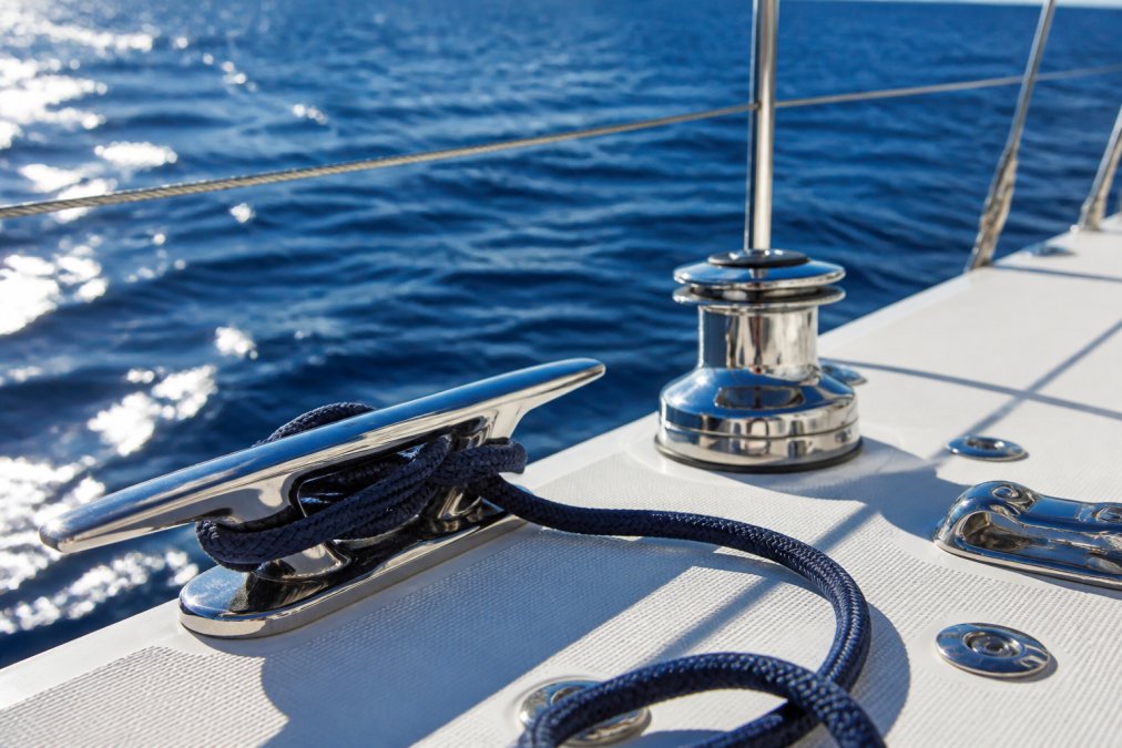 Close-up of polished 316 stainless steel cleats and deck hardware mounted on a sailboat deck, with deep-blue ocean water visible in the background under bright afternoon sunlight