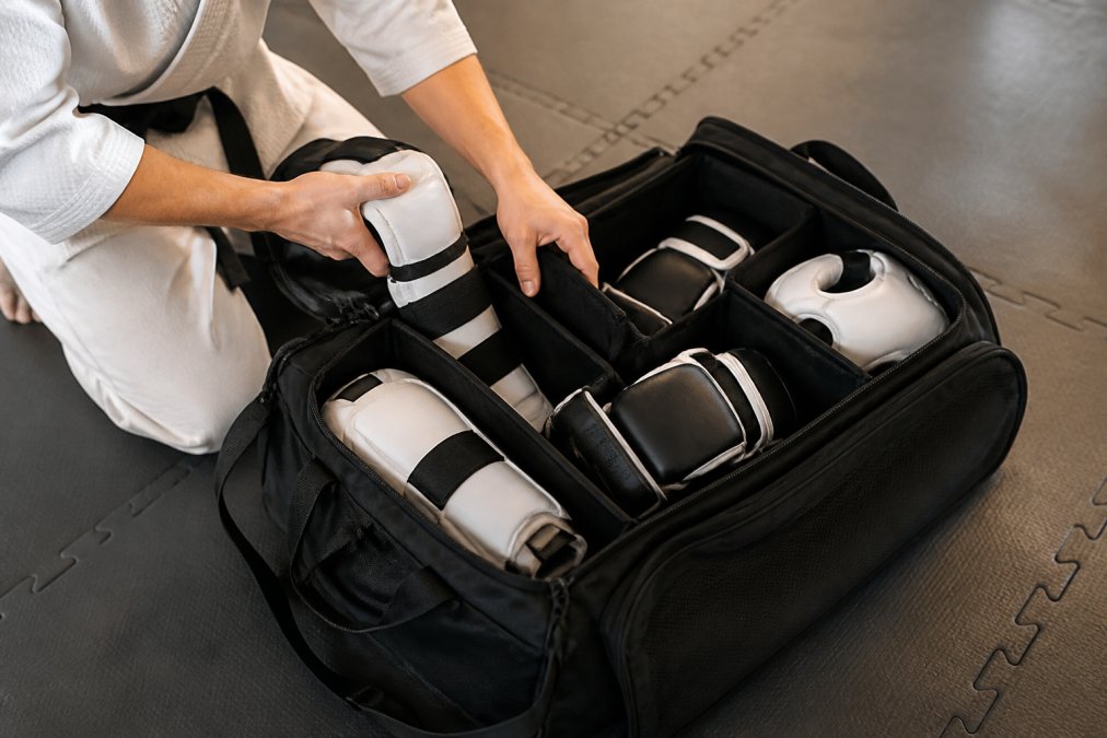 Martial artist organizing sparring gear including shin guards, boxing gloves, and protective pads inside an open duffel bag with multiple compartments, showing organized storage and compartmentalization