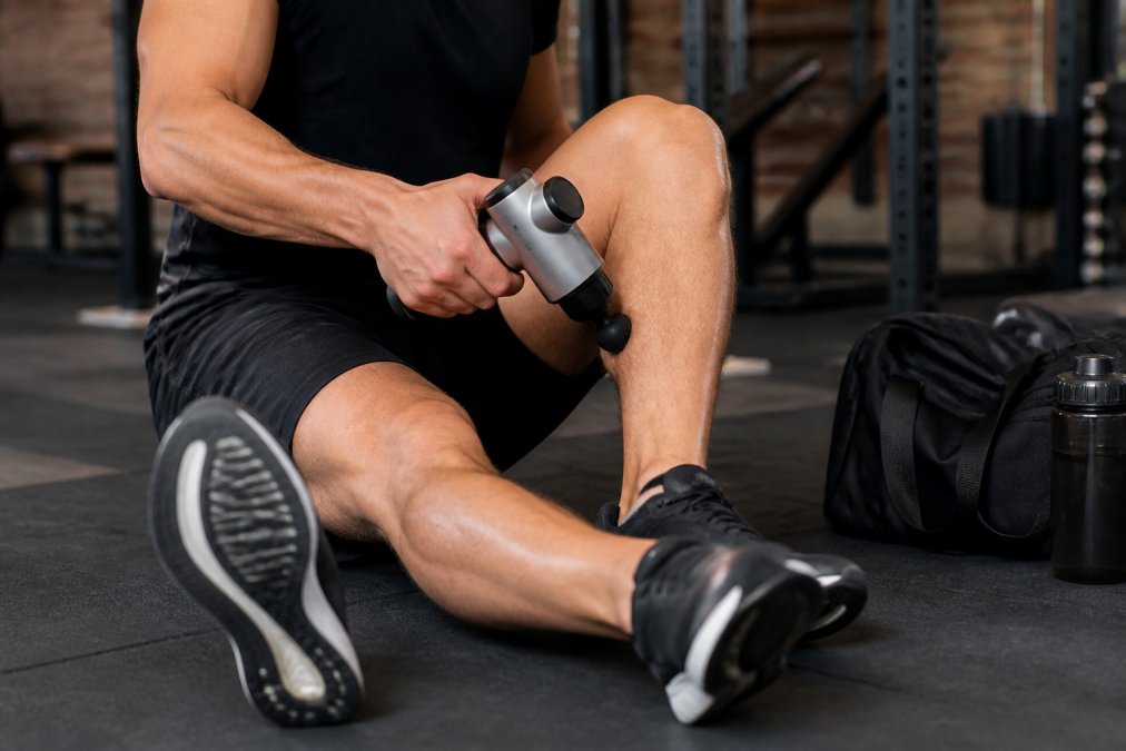 Close-up of a person applying a handheld massage device to their calf muscle post-workout in a gym setting, gym bag and water bottle visible in the background, natural athletic lighting