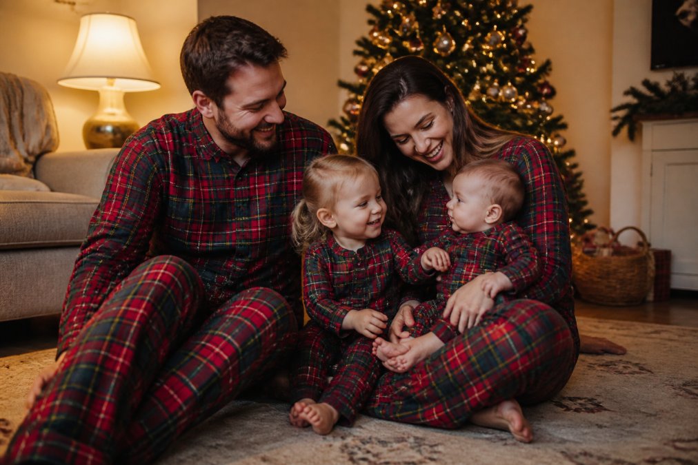A cheerful family of four, two parents, a toddler, and a baby, wearing coordinating red and green plaid flannel pajamas, seated on a living room floor near a lit Christmas tree, laughing naturally in warm lamplight