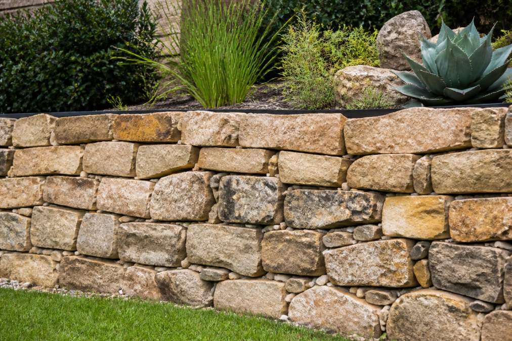 Finished natural stone retaining wall in a residential backyard landscape, showing varied warm tan and gray stone colors with natural gaps between stones allowing water drainage, backed by landscaping plants