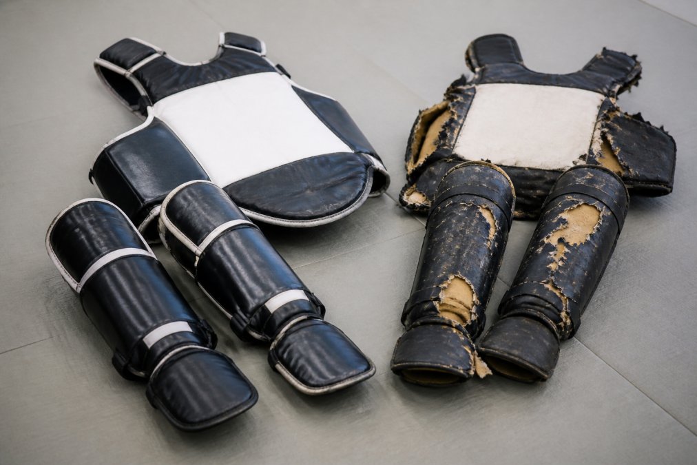 Close-up of well-worn but intact martial arts shin guards and chest protector laid out on a dojo mat next to a worn-out torn pair, showing the contrast in material quality and construction