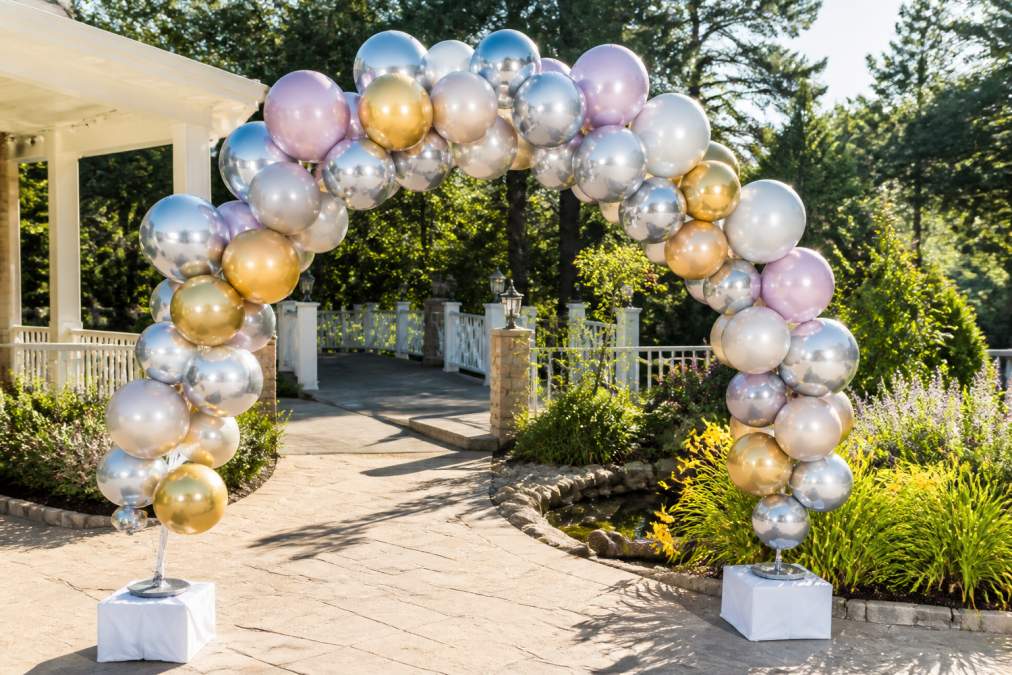 Professional outdoor balloon arch installation in bright sunlight, showing UV-resistant foil balloons anchored with weighted bases at a garden venue