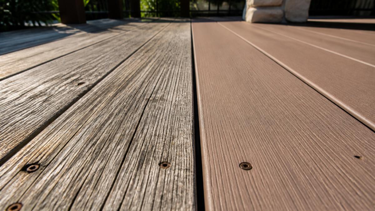 A close-up photograph of a weathered gray wooden deck board beside a clean composite deck board, sunlight raking across both surfaces to show grain texture and surface wear