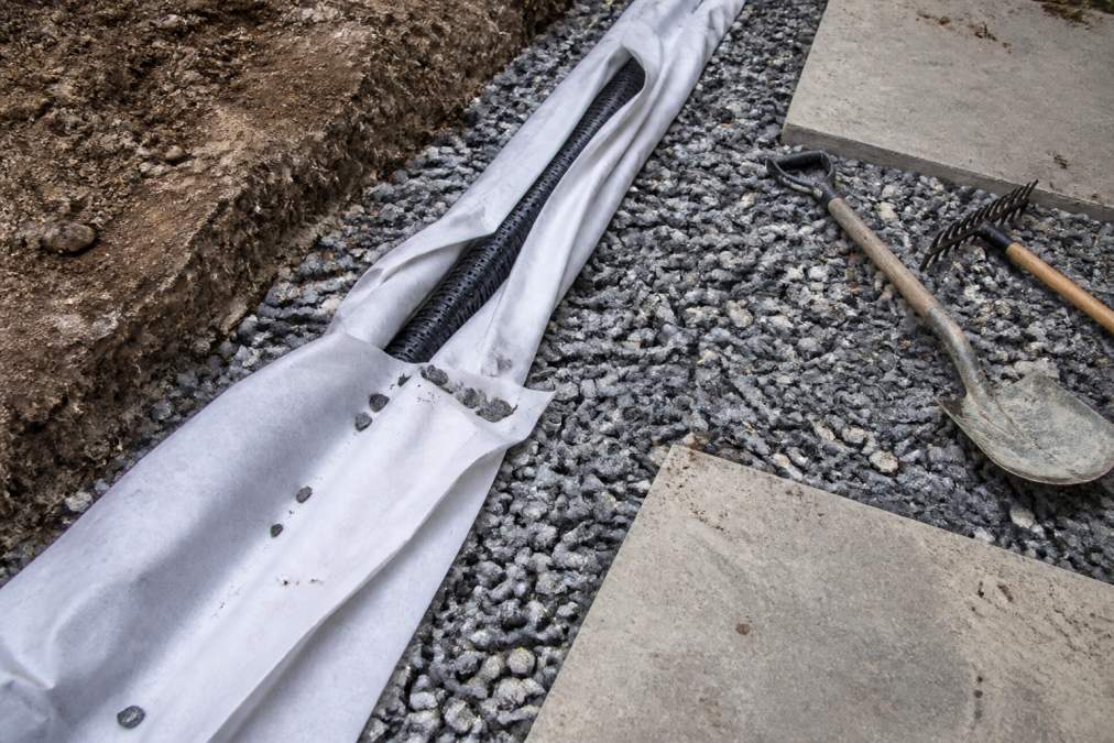 Overhead view of a patio installation in progress showing perforated drain pipe wrapped in geotextile fabric and surrounded by angular crushed stone base material, with installation tools visible