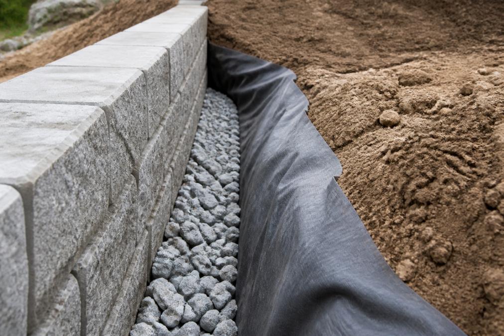 Close-up of installed concrete retaining wall blocks with visible drainage gravel and geotextile fabric layered behind the wall, showing proper construction detail with crushed limestone visible