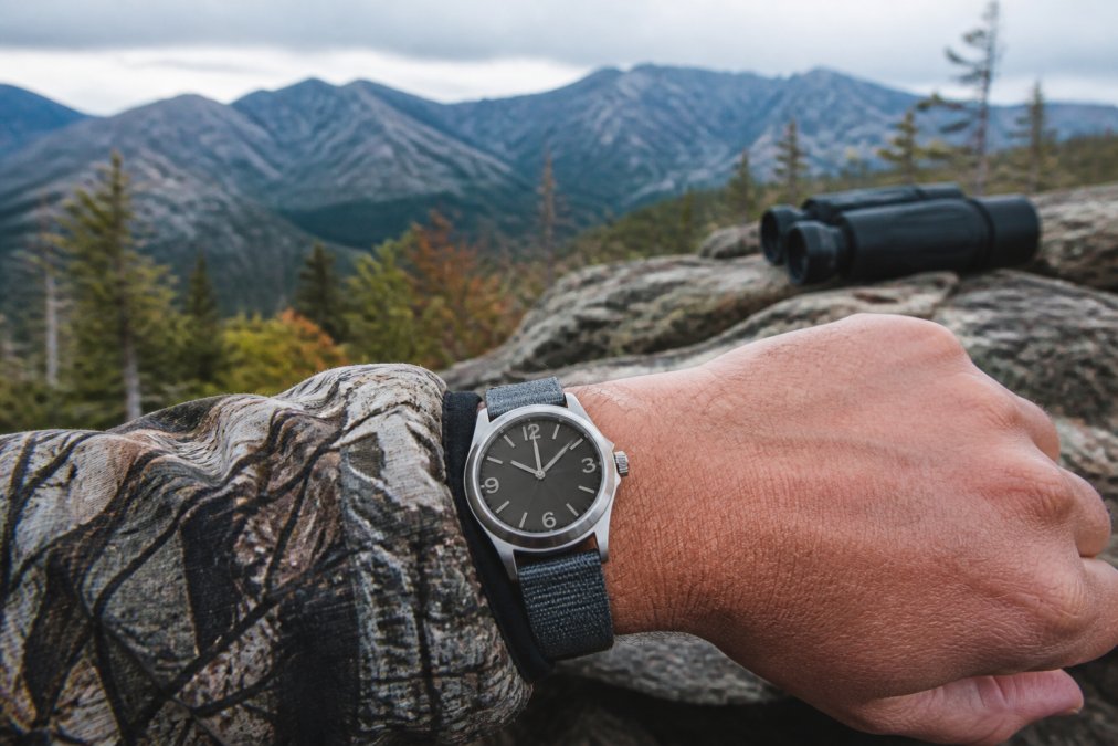 Mechanical hunting watch displayed on hunter's wrist in backcountry setting with forest terrain and natural outdoor lighting visible