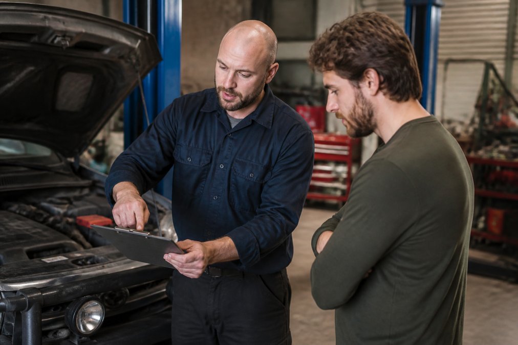 Mechanic discussing vehicle repairs with a customer in a modern workshop, pointing at diagnostic results, showing transparency and professional communication with warm lighting