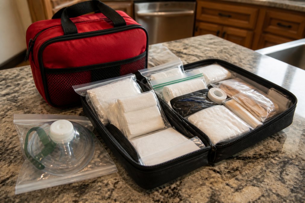 A home first aid kit laid open on a kitchen counter, showing neatly organized bandages, gauze pads, antiseptic wipes, and a CPR mask in clear resealable bags, with a red medical bag nearby
